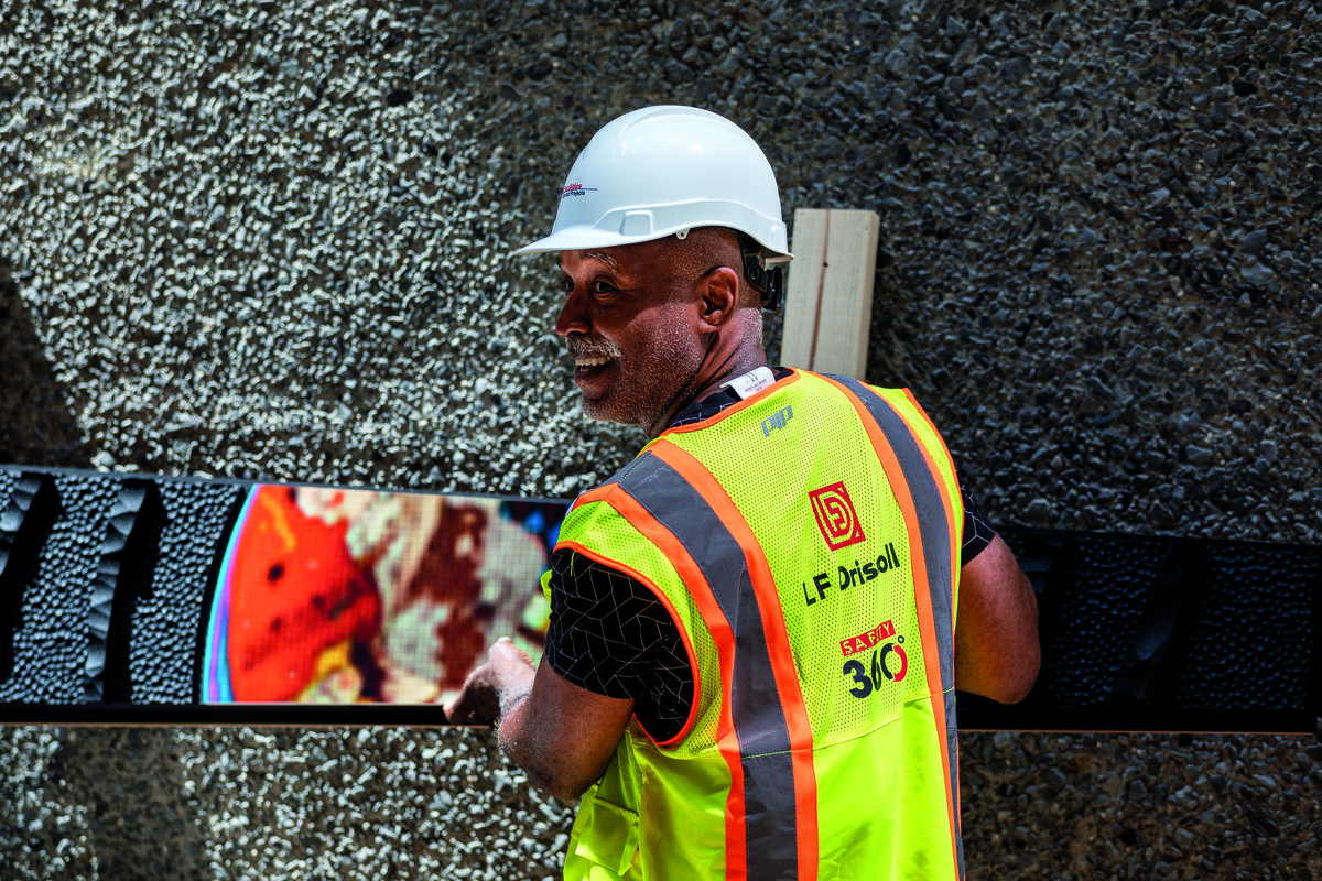 A man wearing a hardhat and construction vest holds up a board with textured black pieces and abstract colorful pattern.