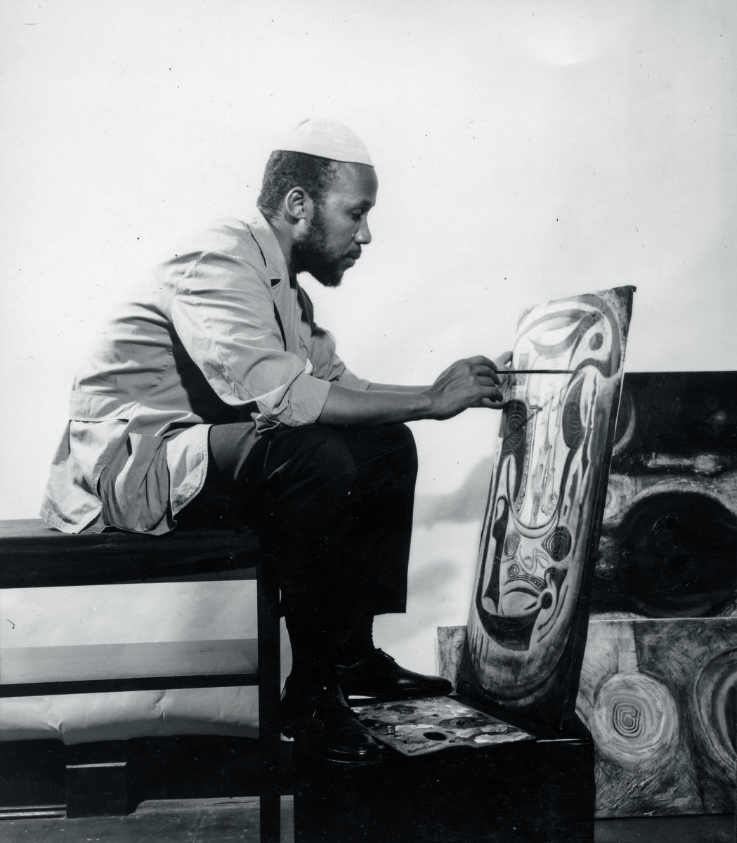 The artist sitting on a low stool painting a canvas.