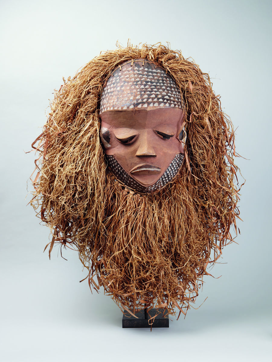 An african mask depicting a face with downturned eyes, and long hair made from a natural material.