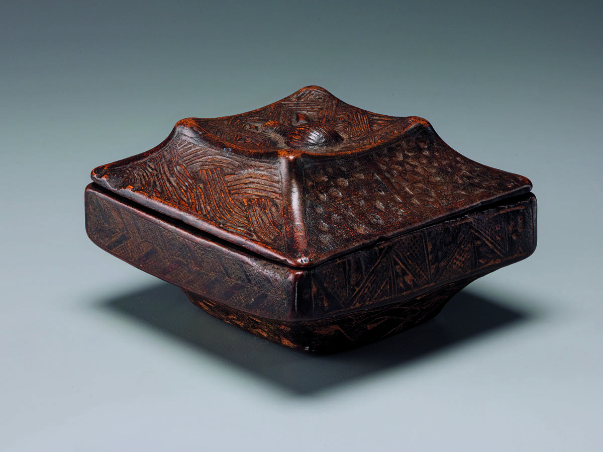 A wooden square box that sits on a built in foot with a tented lid with abstract carvings.